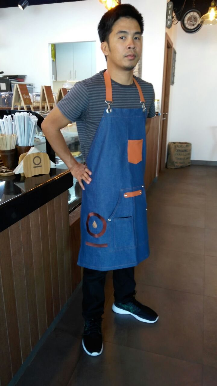 Barista standing in a café wearing a denim bib apron with tan leather straps over a grey T-shirt