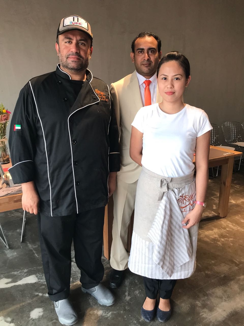 Restaurant team in uniform: a chef in a black chef jacket and cap, a manager in a beige suit and a server in a white T-shirt with a striped waist apron