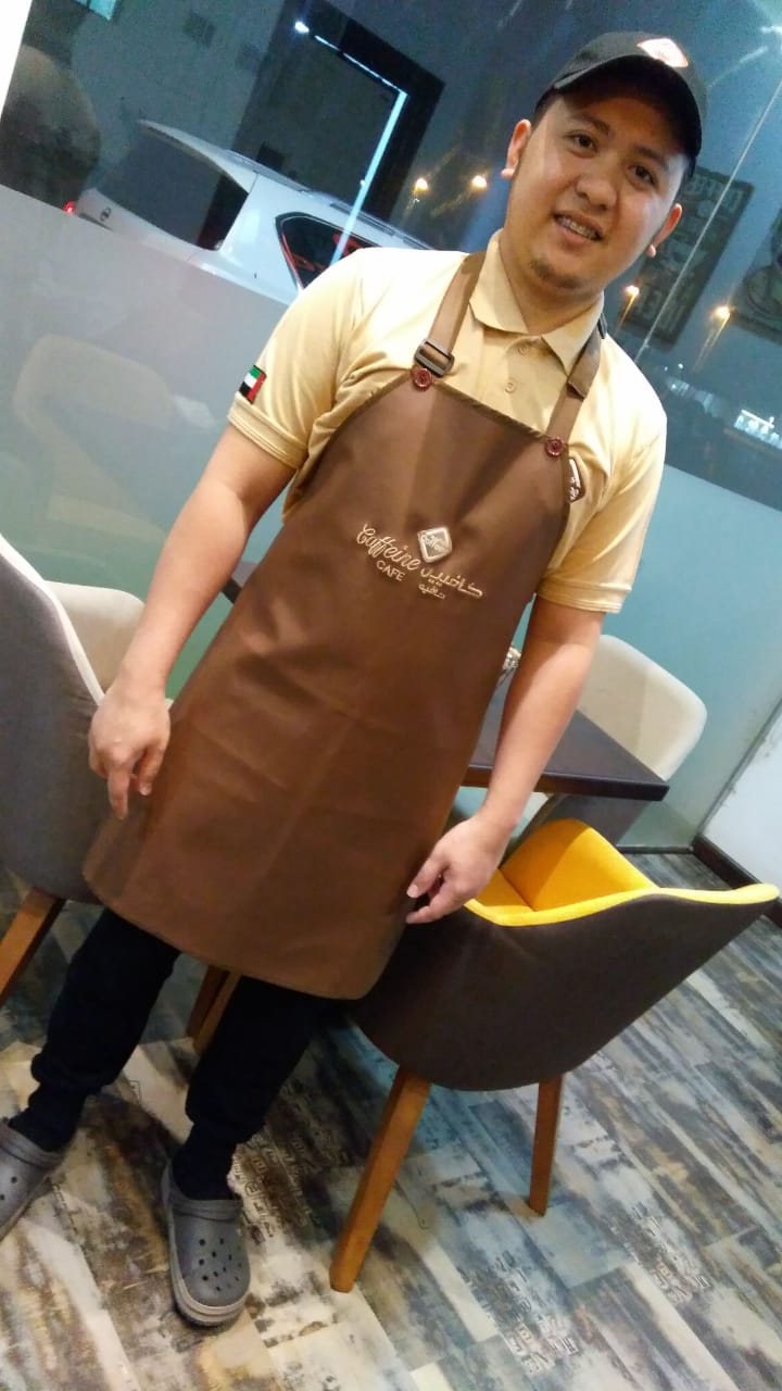 Café worker wearing a brown bib apron with an embroidered logo over a beige T-shirt