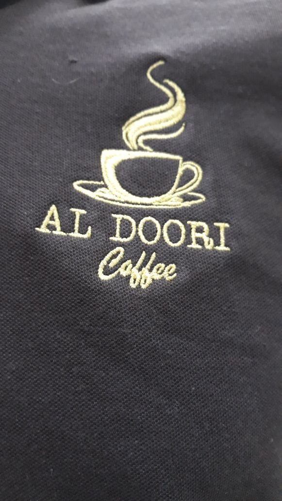 Close-up of gold-thread embroidery reading 'Al Doori Coffee' with a steaming cup motif on a grey apron