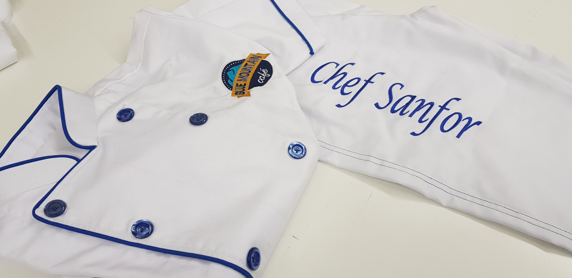 Close-up of two white chef jackets with blue piping and buttons, one embroidered 'Chef Sanfor' in blue script and a round café badge
