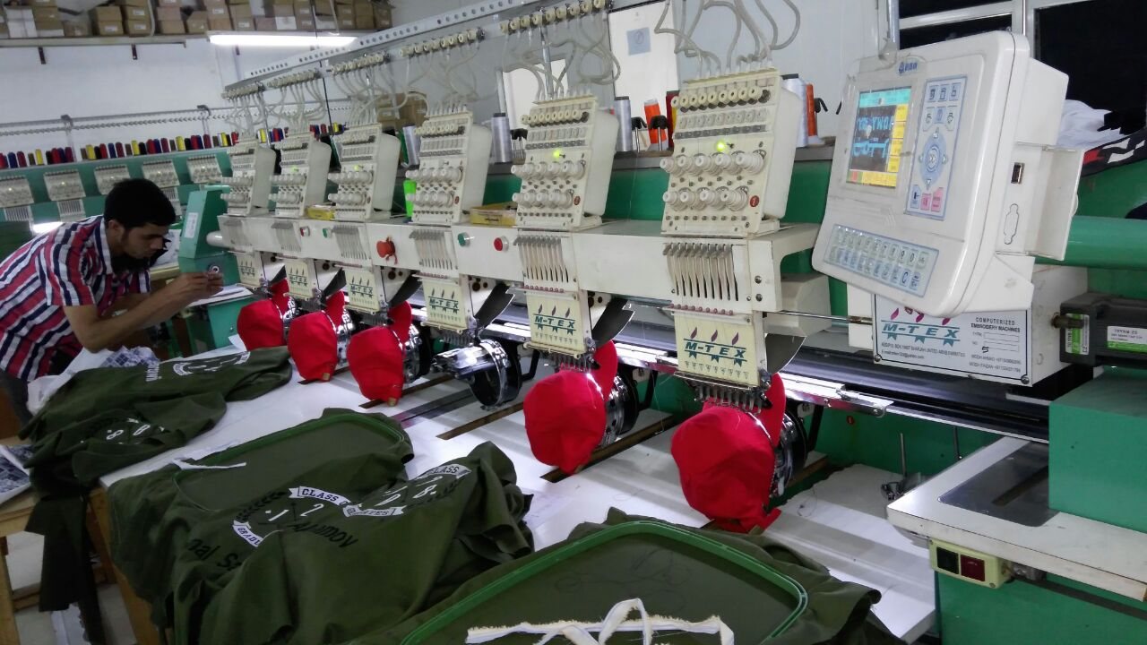 Embroidery workshop: a technician tends a multi-head embroidery machine stitching logos onto red caps, with green garments laid out in front